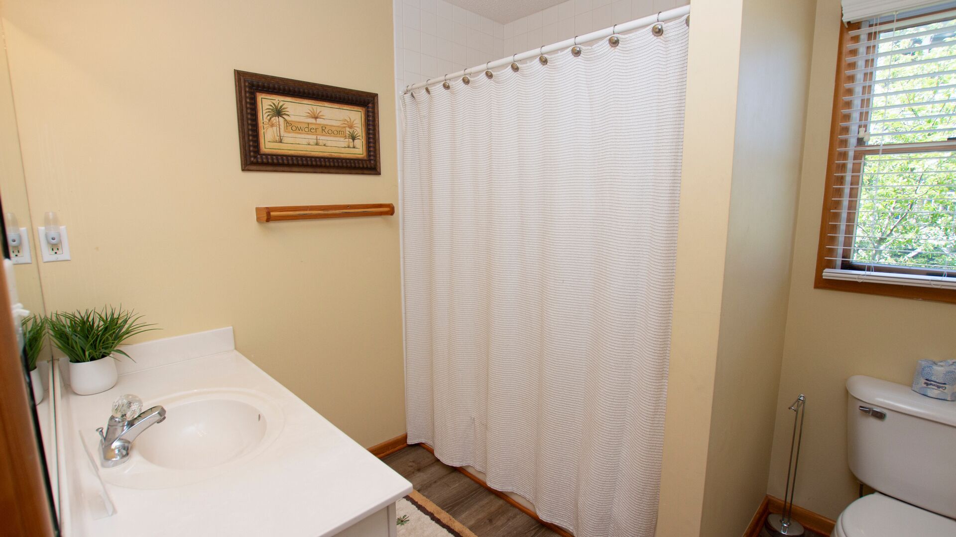 A bathroom with a shower curtain, a sink, a toilet, a framed sign on the wall, and a window with blinds.
