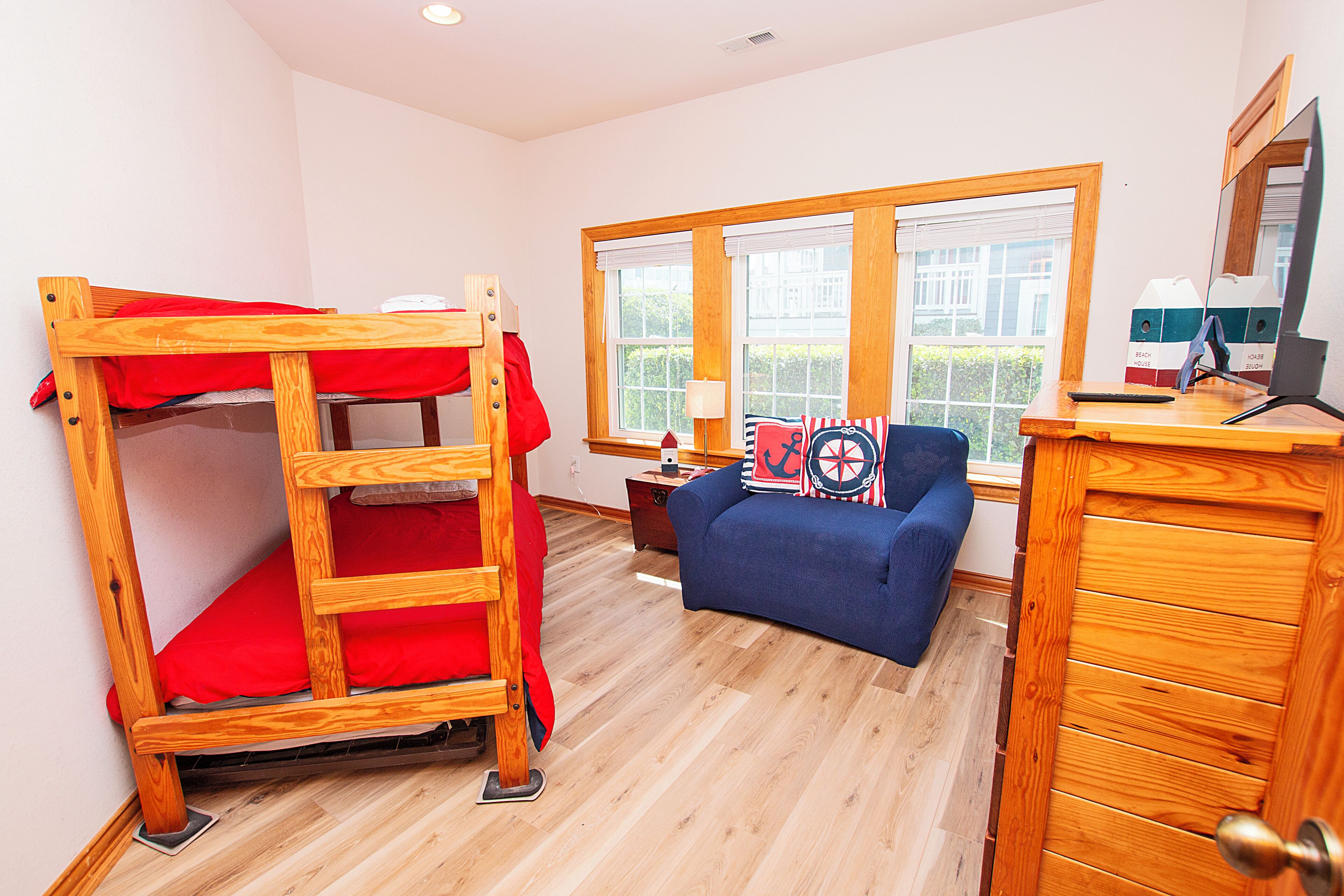 A bunk bed with red bedding, a blue sofa with nautical-themed pillows, a wooden desk with a computer, and a window with blinds.