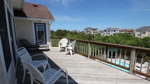 Um deck de madeira com cadeiras de descanso, uma pequena mesa e vista para uma piscina e casas vizinhas.