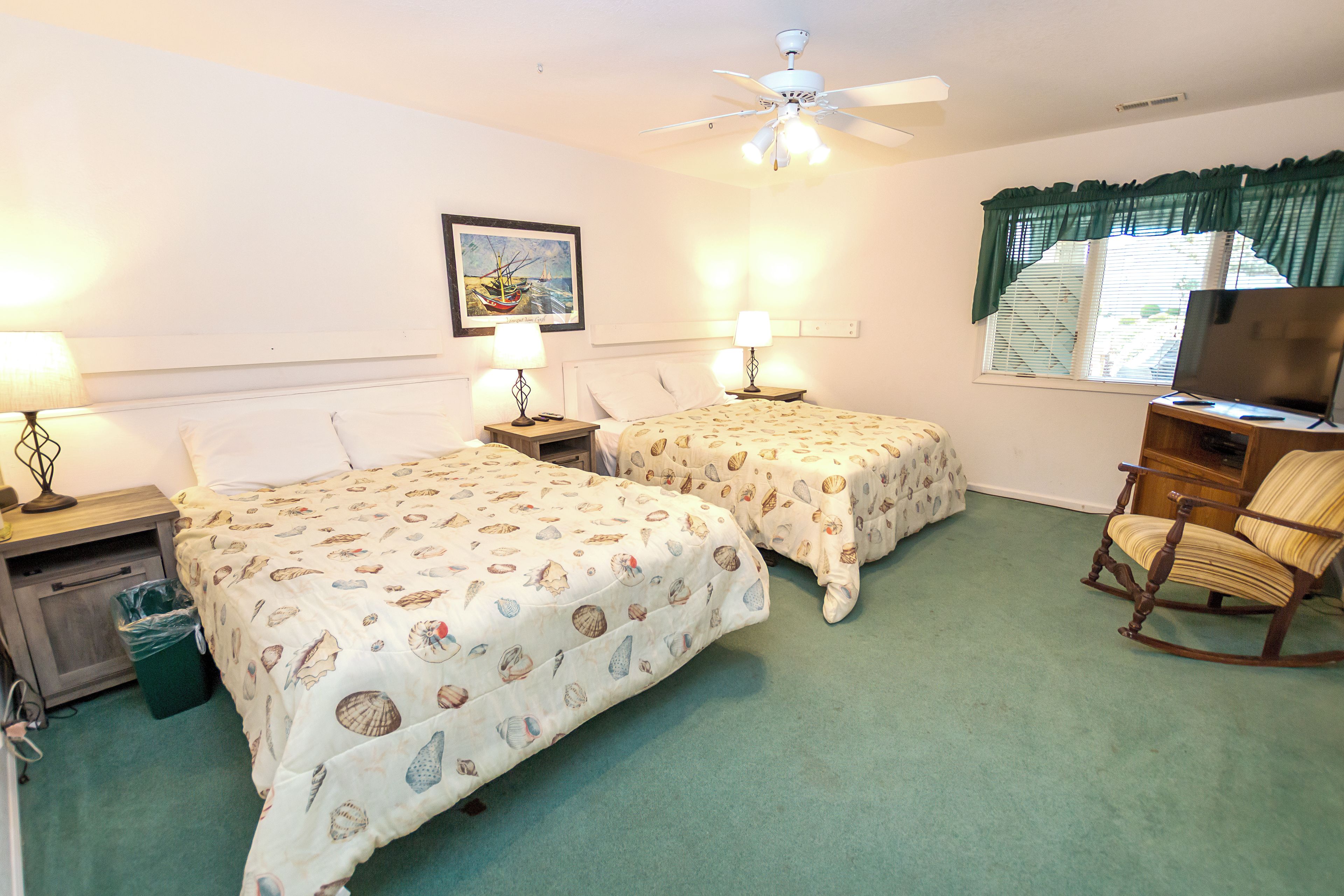 A hotel room with two beds, a ceiling fan, a painting, a television, and a rocking chair.