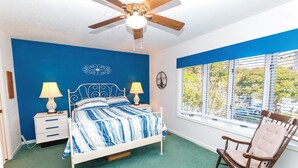A bedroom with a bed, two bedside tables with lamps, a ceiling fan, a rocking chair, a window with blinds, and a wall clock.
