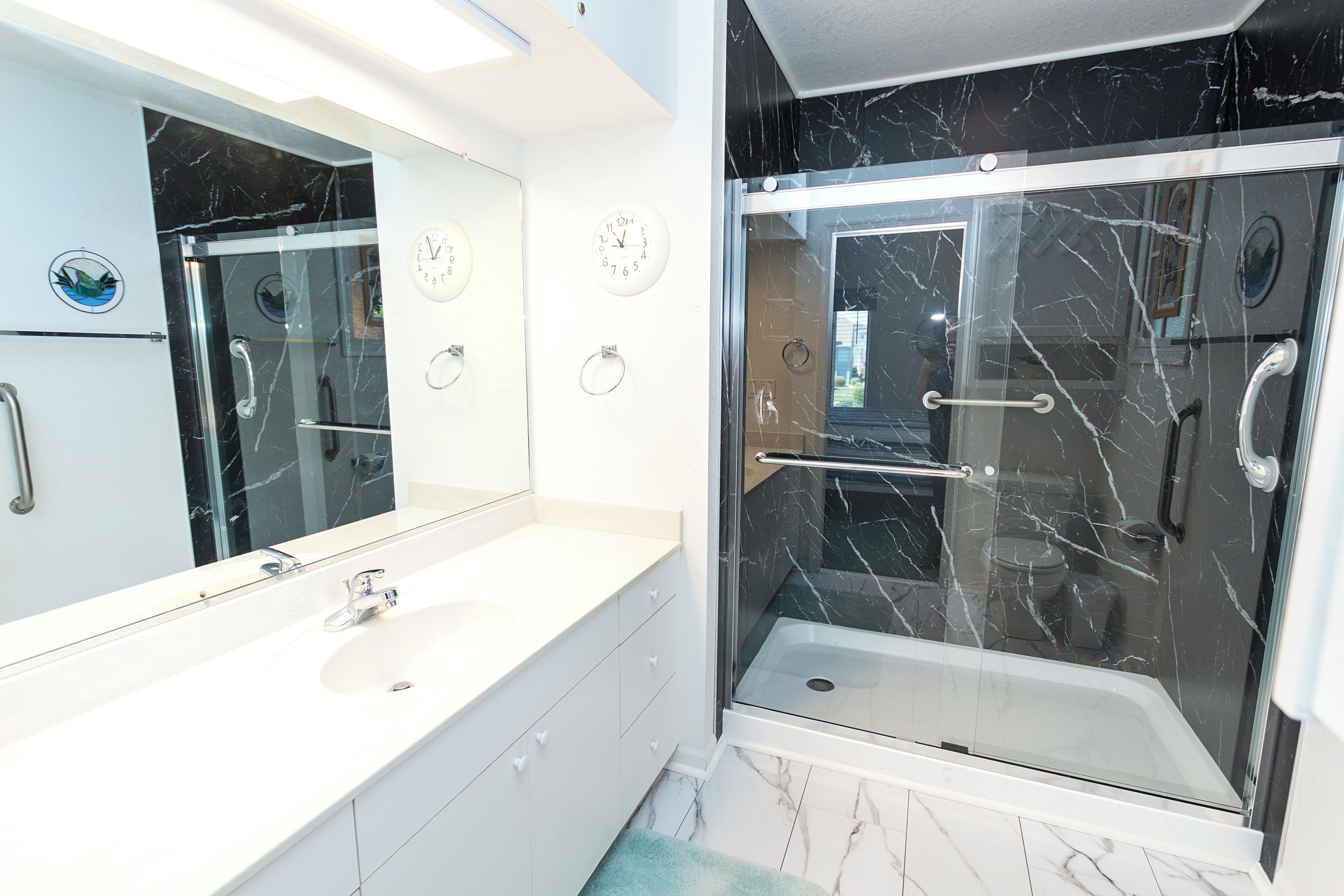 Un baño con lavamanos blanco, espejo, reloj y cabina de ducha con paredes de vidrio.