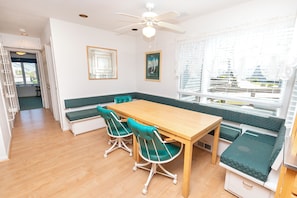 Quarto com mesa de madeira, assentos acolchoados em tons de verde, ventoinha no teto e janela com vista para prédios.