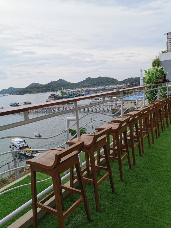 A waterfront bar with wooden stools and a view of boats and a pier. - Flamingo Avia Labuan Bajo (Labuan Bajo)