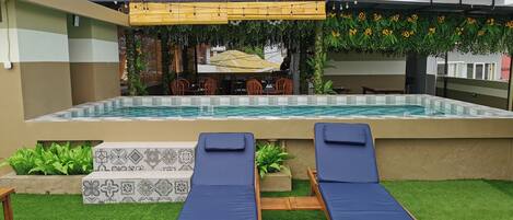 A pool area with two lounge chairs, a covered seating area, and a restaurant in the background.