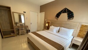 Quarto de hotel com uma cama grande, cabeceira de madeira, mesa de cabeceira com abajur, pequena mesa com uma cadeira, espelho e guarda-roupa.