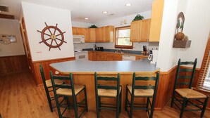 A kitchen with wooden cabinets, a central island with bar stools, and a ship's wheel decoration on the wall. - Oceanside, Private Pool, Hot Tub, Sun Decks, WC1077, Seaside Manor (Corolla)