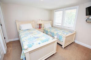 A bedroom with two single beds, a nightstand with a lamp, a window with blinds, and a TV mounted on the wall. - Oceanside, Community Amenities, Prv Pool, Rec Rm, 5, Carolina on my Mind (Corolla)