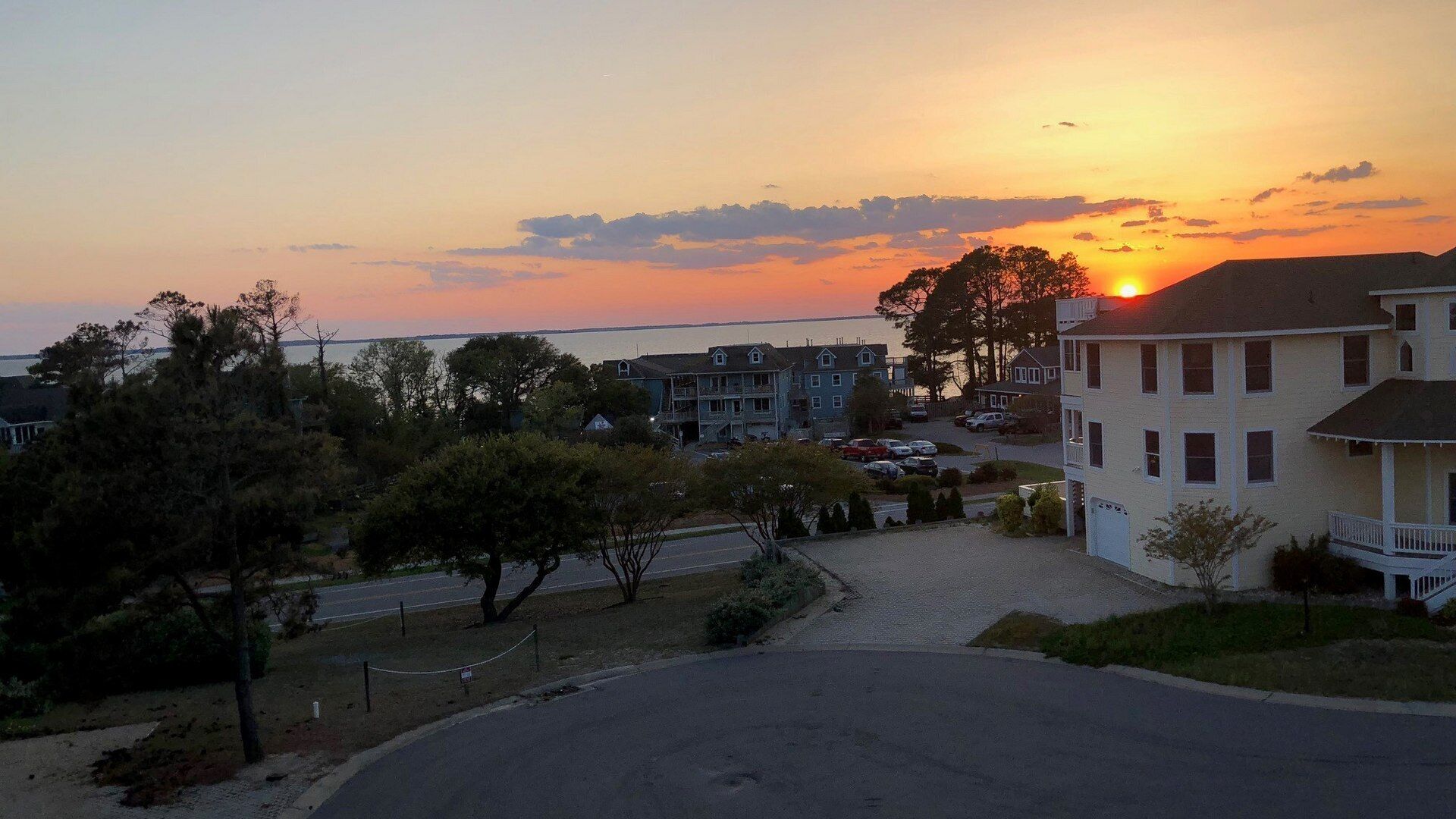 A residential area with a sunset view, featuring multi-story buildings, a parking lot, and a road leading to the beach.