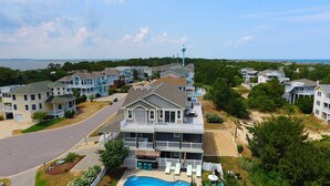 A coastal residential area with a large house featuring a swimming pool, multiple balconies, and a lighthouse in the background. - Oceanside, Priv Pool, ELEV, Hot Tub, Keyless entry, BH3, Knot 4 Sail (Corolla)