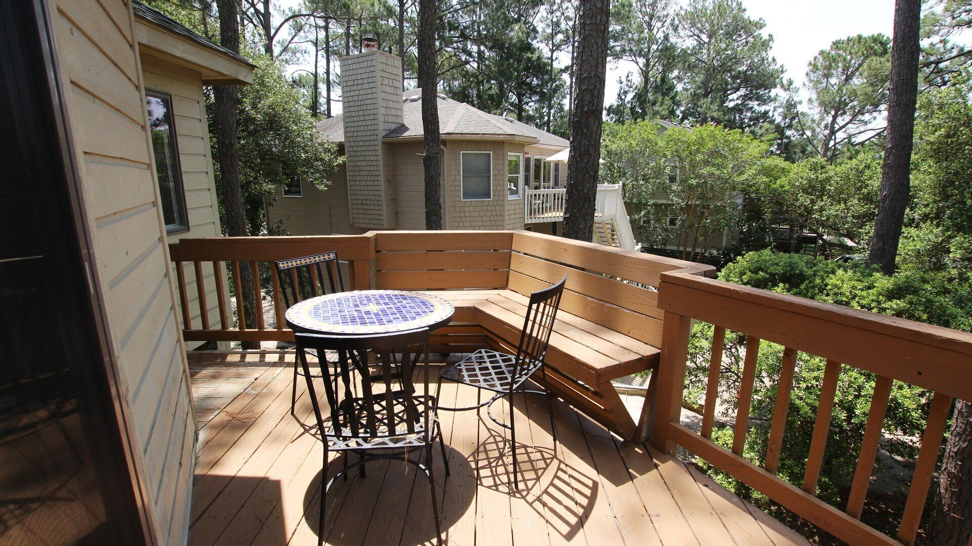 A wooden deck with a round table and chairs, a bench, and a view of a house surrounded by trees.