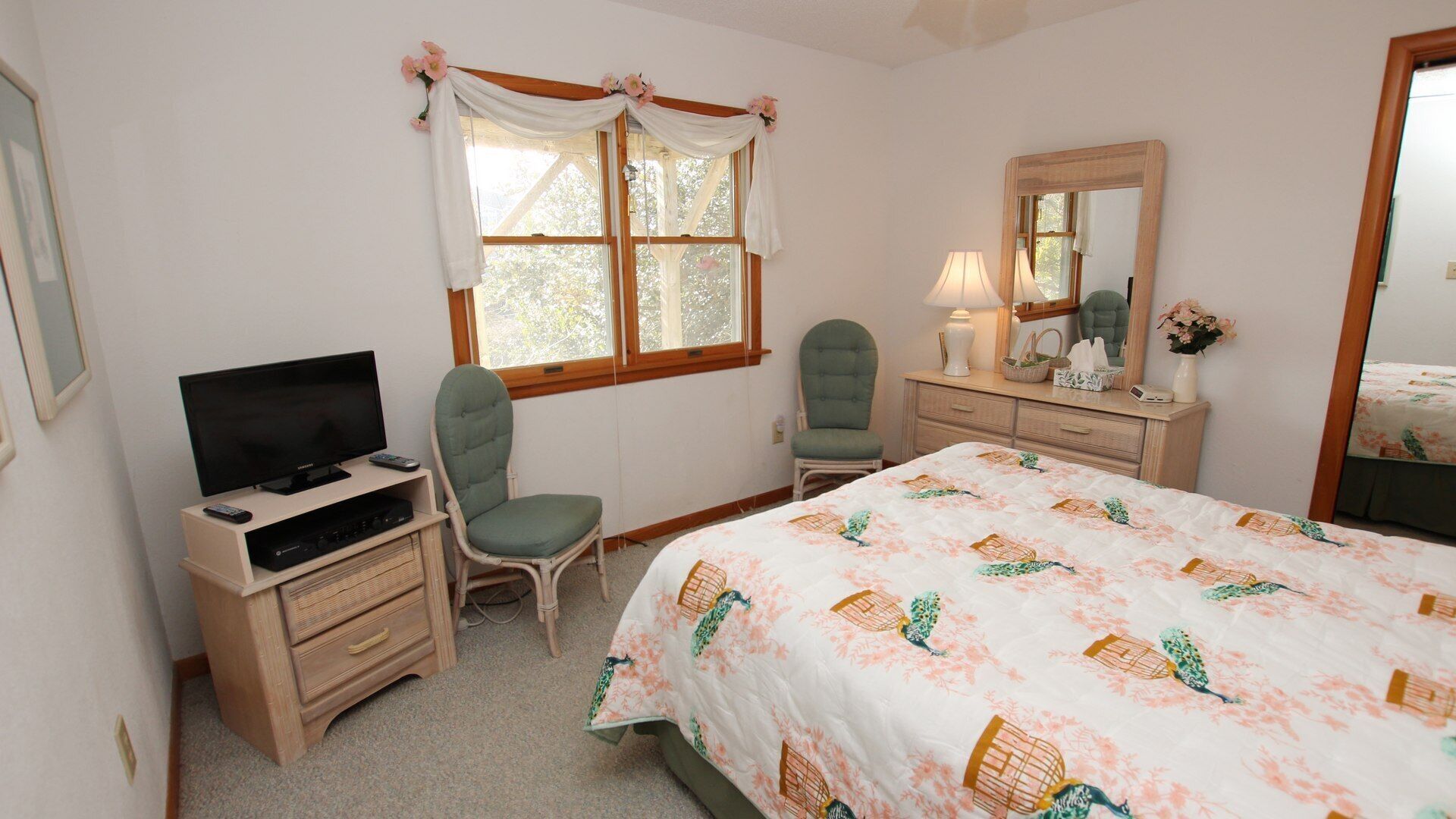 A bedroom with a bed, two chairs, a TV stand, a dresser, a mirror, and a window with curtains.
