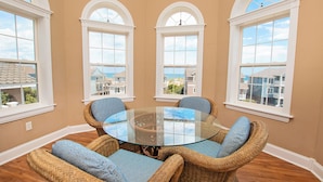 A room with a glass-top table surrounded by wicker chairs with blue cushions, large windows with white frames, and a view of houses through the windows.