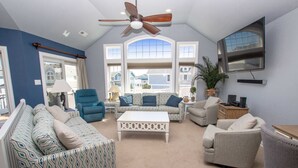 A living room with a ceiling fan, large windows, a flat-screen TV, and a variety of seating options including patterned sofas and armchairs. - Oceanside, Hot Tub, Dogs Welcome, Community Pool, BU13B, Tropical Dreams B (Corolla)