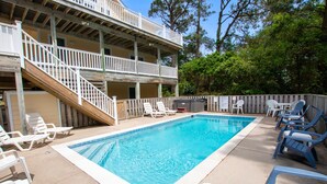 Un espace piscine doté d’une terrasse en bois, de chaises longues blanches et bleues, et d’un bâtiment à deux étages avec un balcon.