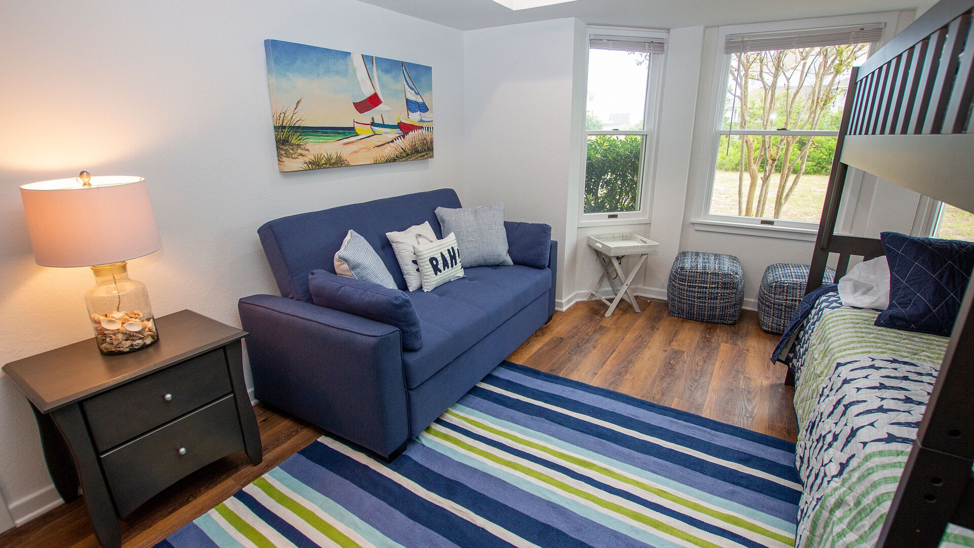 A room with a blue sofa, a small table, a lamp, a striped rug, and a bunk bed.