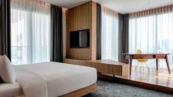 A modern hotel room with a large bed, a flat-screen TV mounted on a wooden wall, a small table with a chair, and large windows with sheer curtains.