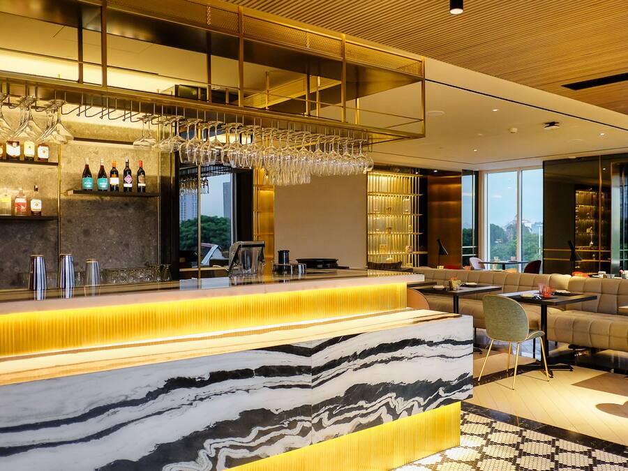 A modern bar area with a marble countertop, wine glasses, and bottles. There is a seating area with sofas and a view of the outdoors.