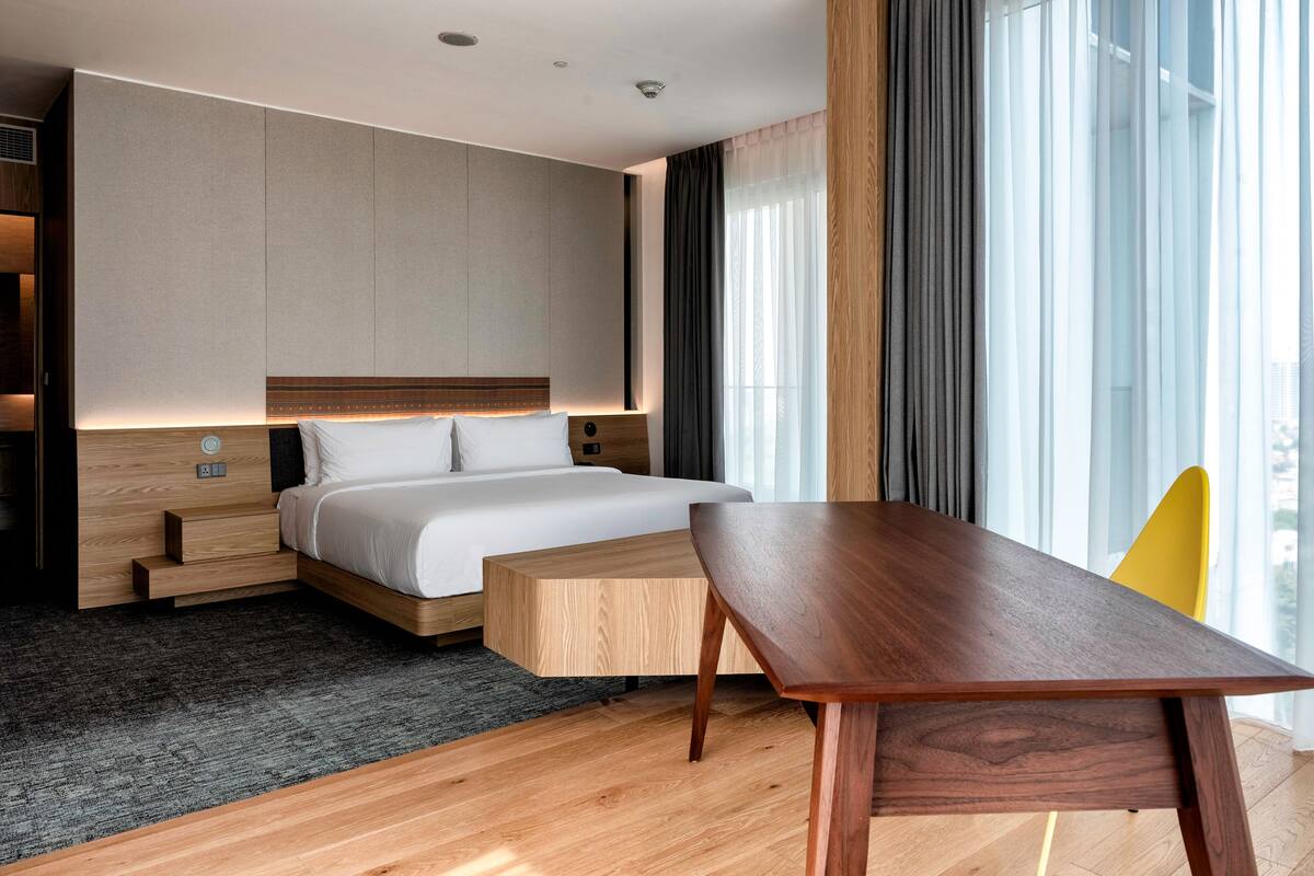 A modern hotel room with a large bed, a wooden nightstand, a desk, and a view of the city through large windows.