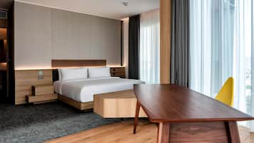 A modern hotel room with a large bed, a wooden nightstand, a desk, and a view of the city through large windows.