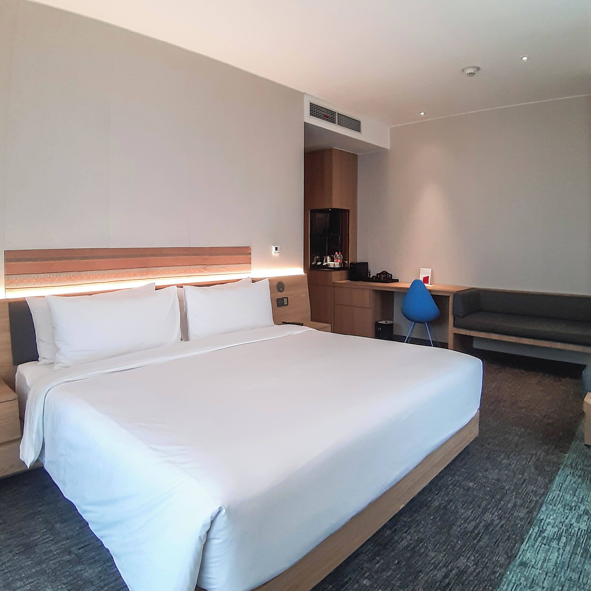A hotel room with a large bed, a desk, a chair, a wardrobe, and a small table with a lamp.