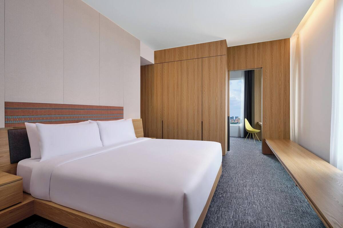 A modern hotel room with a neatly made bed, wooden wardrobe, and a small seating area with a chair.