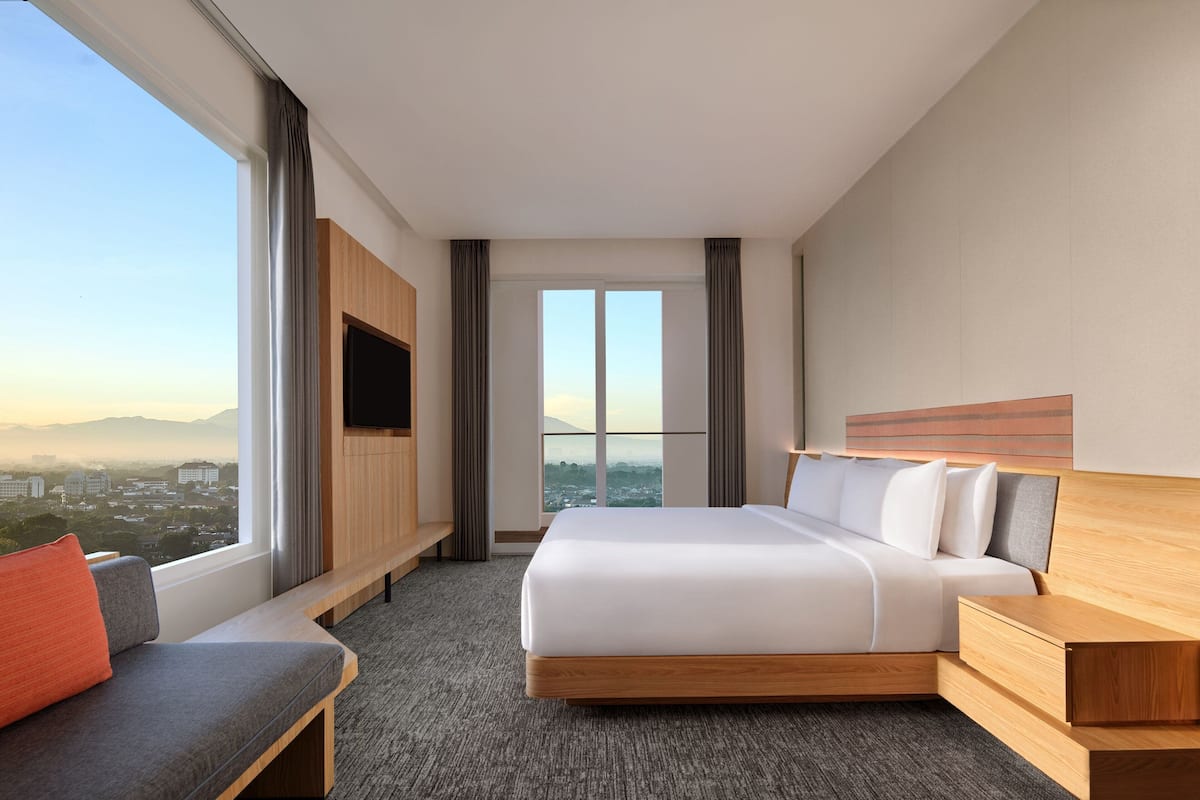 A modern hotel room with a large bed, a seating area, a TV, and a view of the city.