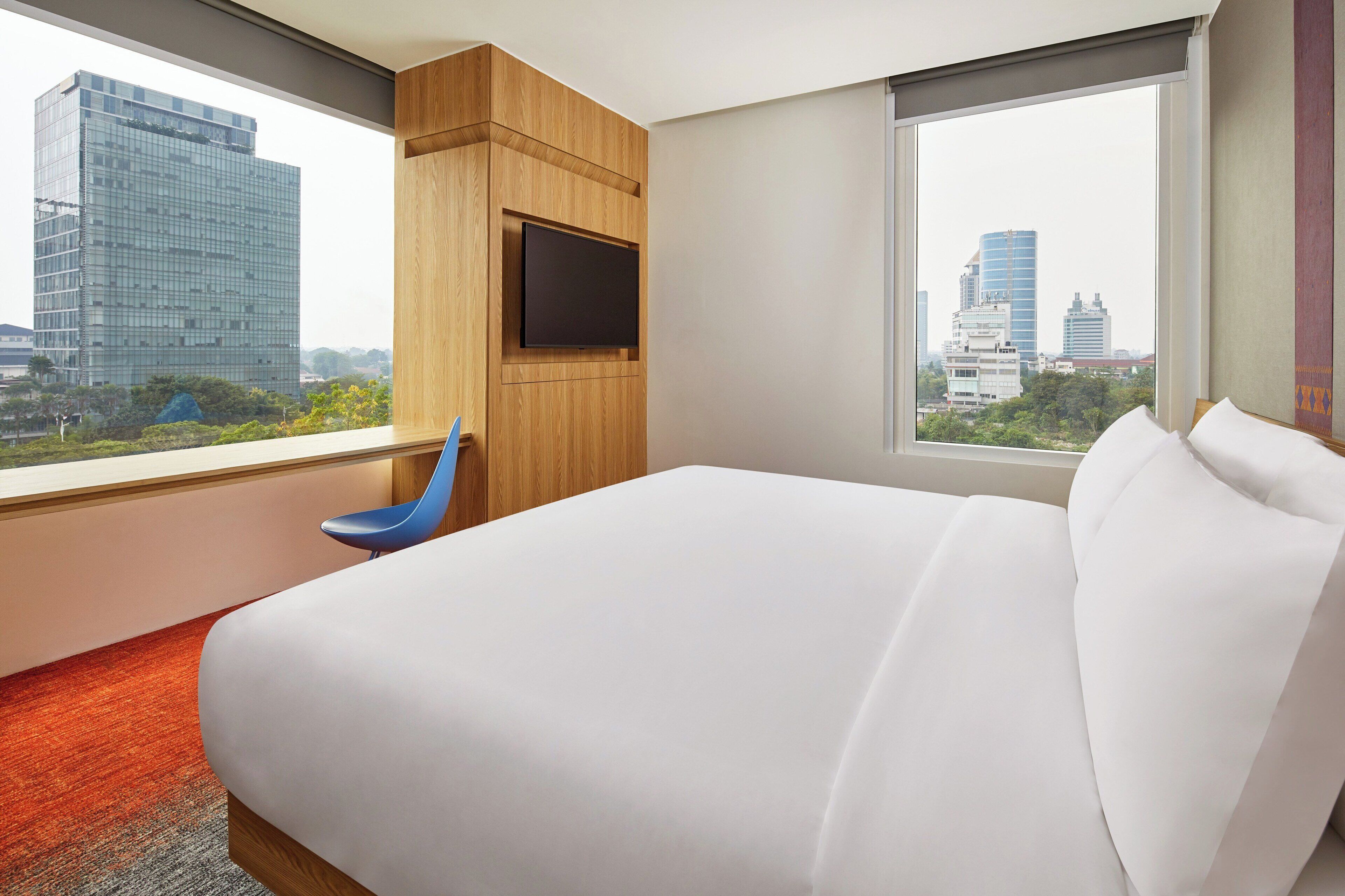 Camera d'albergo con un letto grande, una TV fissata a un pannello di legno e vista sugli edifici della città dalla finestra.
