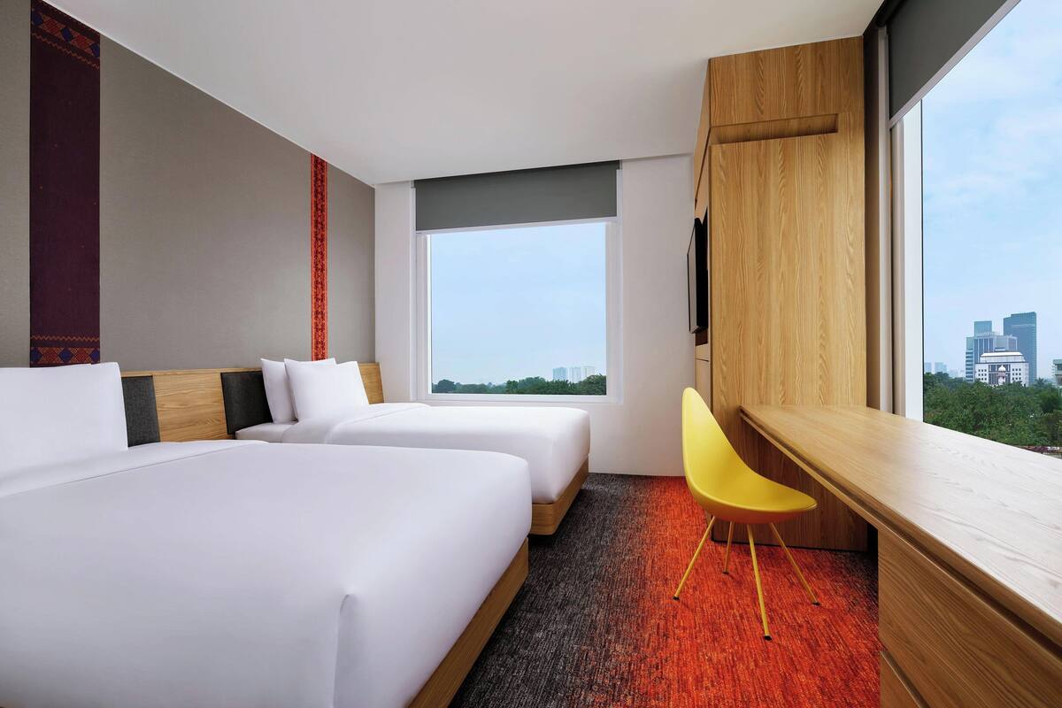 A hotel room with two beds, a desk, a chair, and a large window offering a city view.