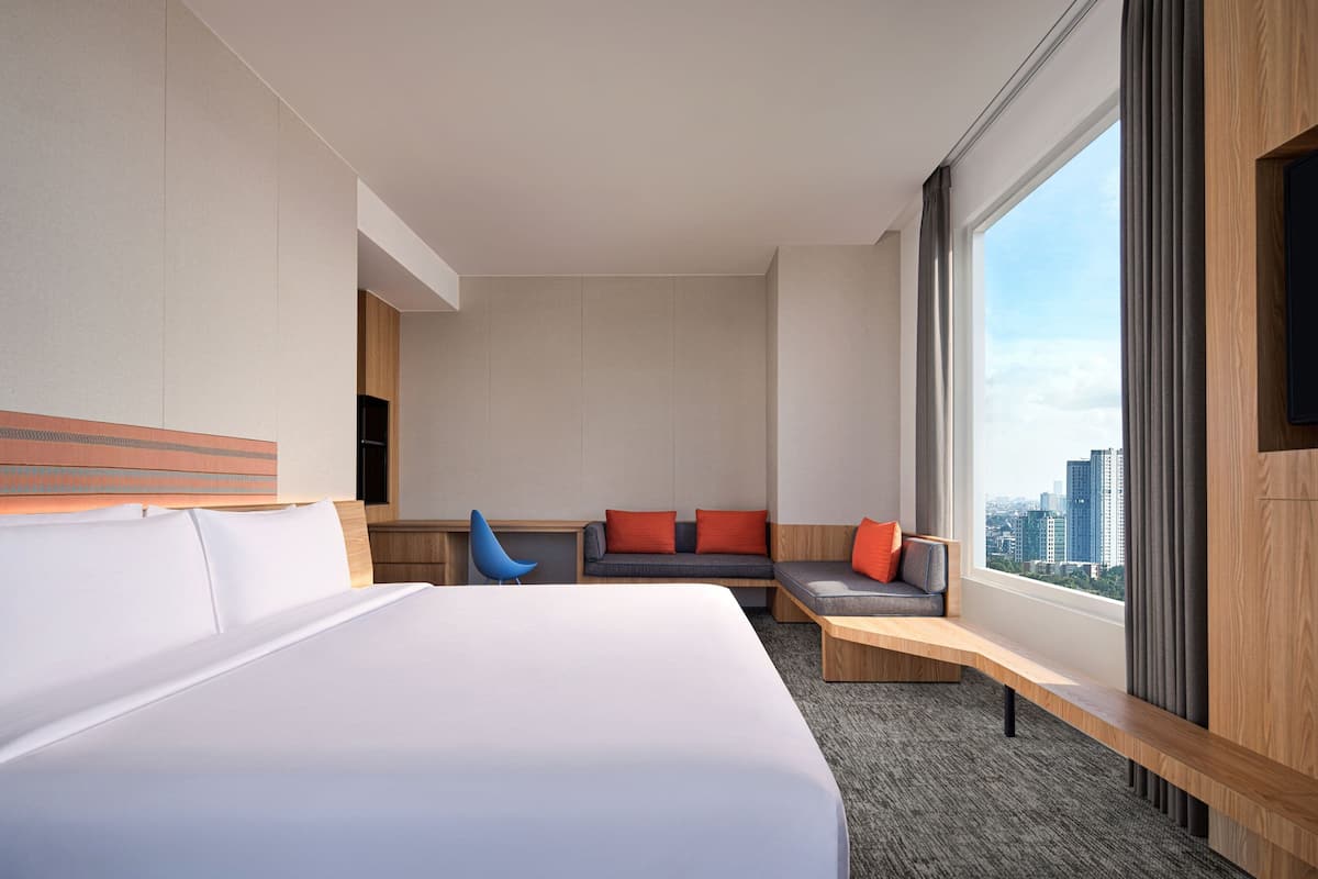 A modern hotel room with a large bed, a seating area with a sofa and chairs, and a large window offering a city view.