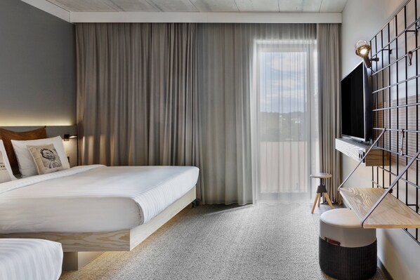 Una habitación de hotel moderna con una cama grande, un televisor montado en una estructura metálica, una mesita auxiliar pequeña y vistas al exterior a través de cortinas translúcidas.