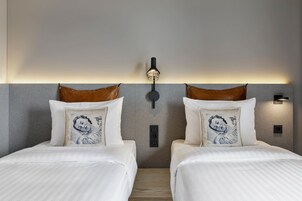Habitación de hotel con dos camas, cada una con un almohadón estampado en azul, una lámpara de pared y una pared gris con textura.