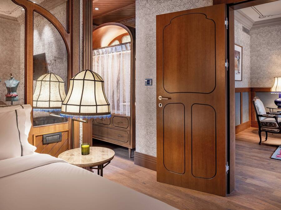 Habitación de hotel con una cama grande, una mesa redonda con lámpara, un armario de madera y vista al baño por un vano arqueado.