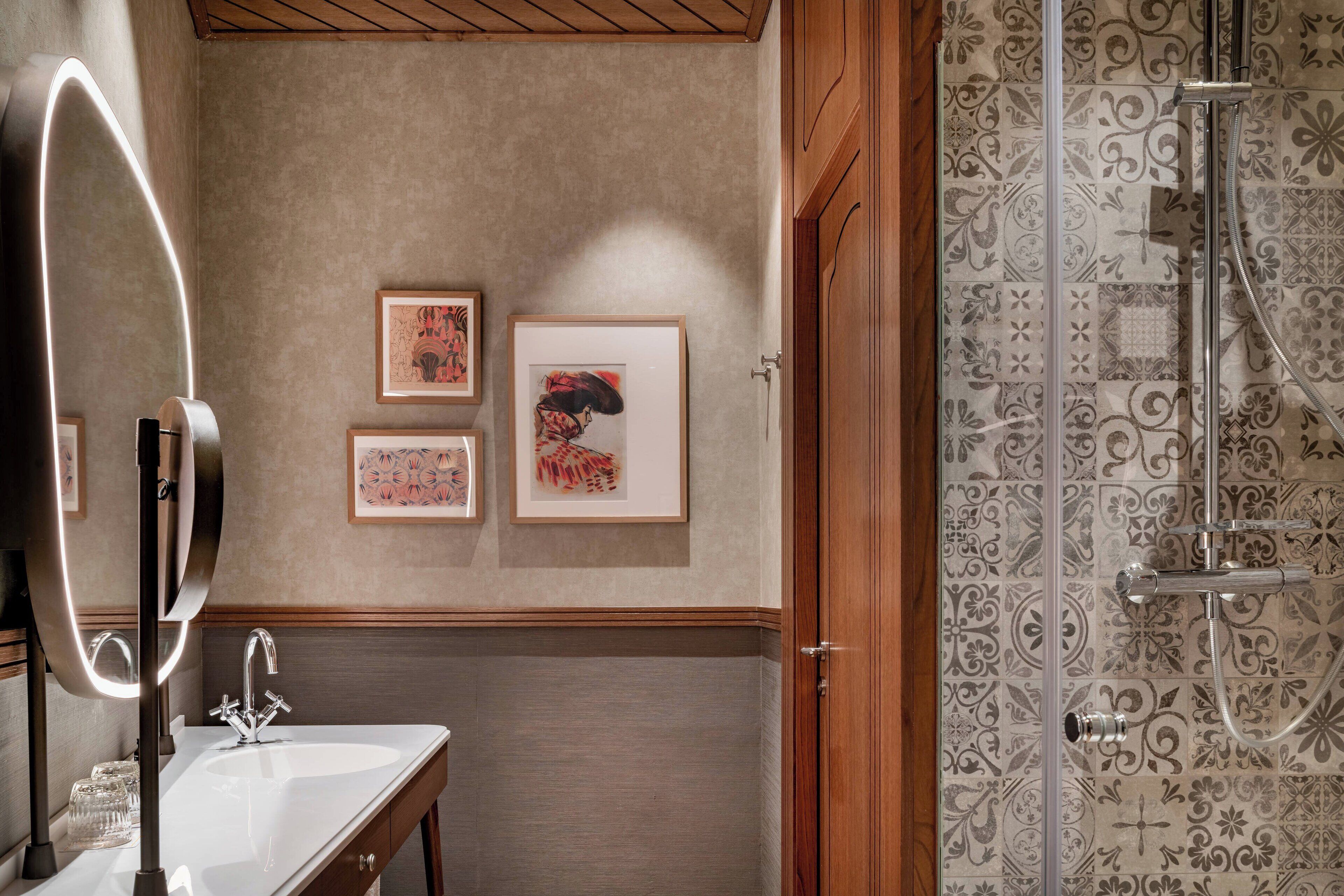 A bathroom with a modern vanity, a large mirror with lights, and a shower with decorative tiles.