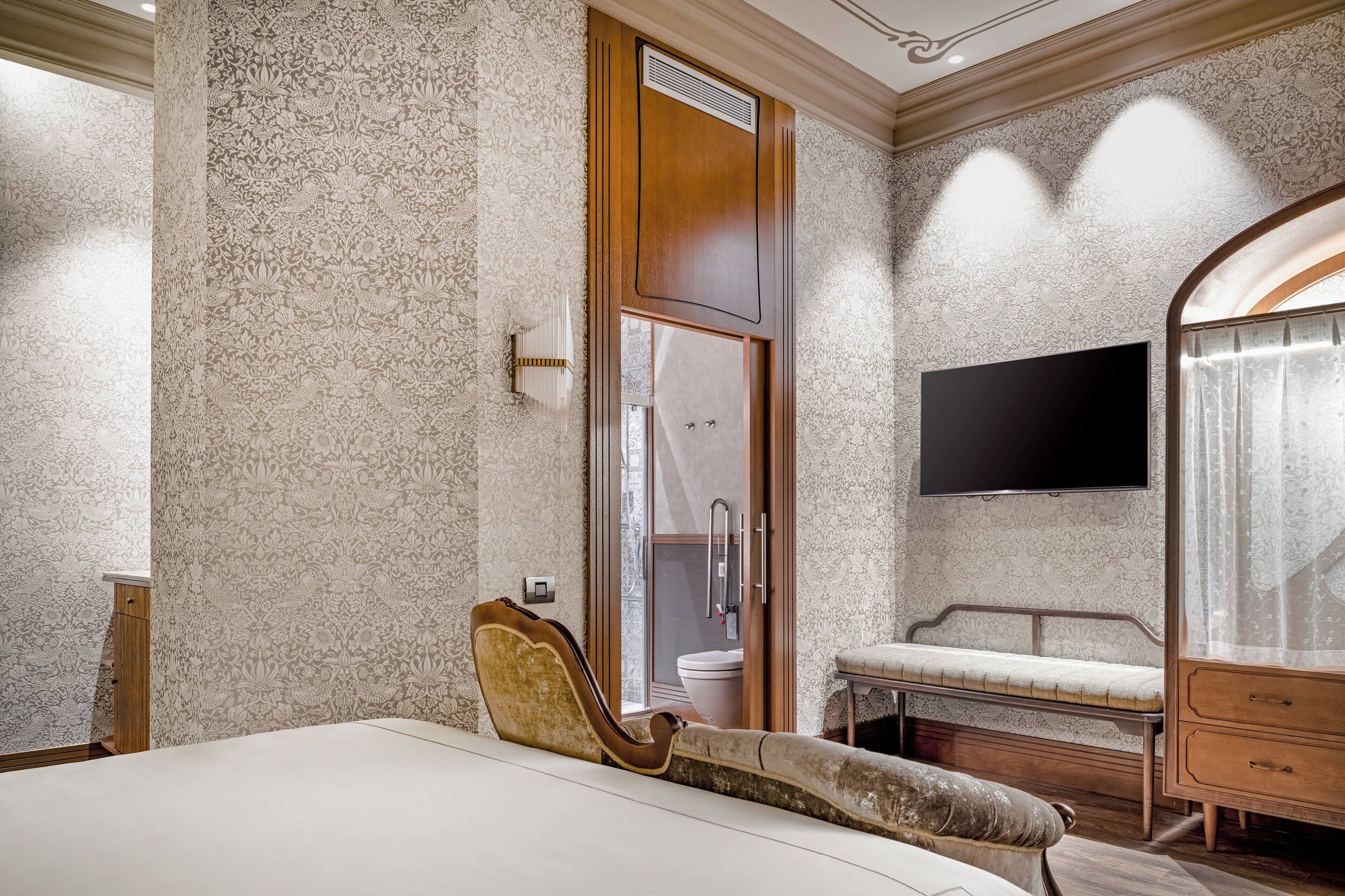 Un dormitorio con cama, un tocador con espejo, un banco y un televisor montado en la pared.