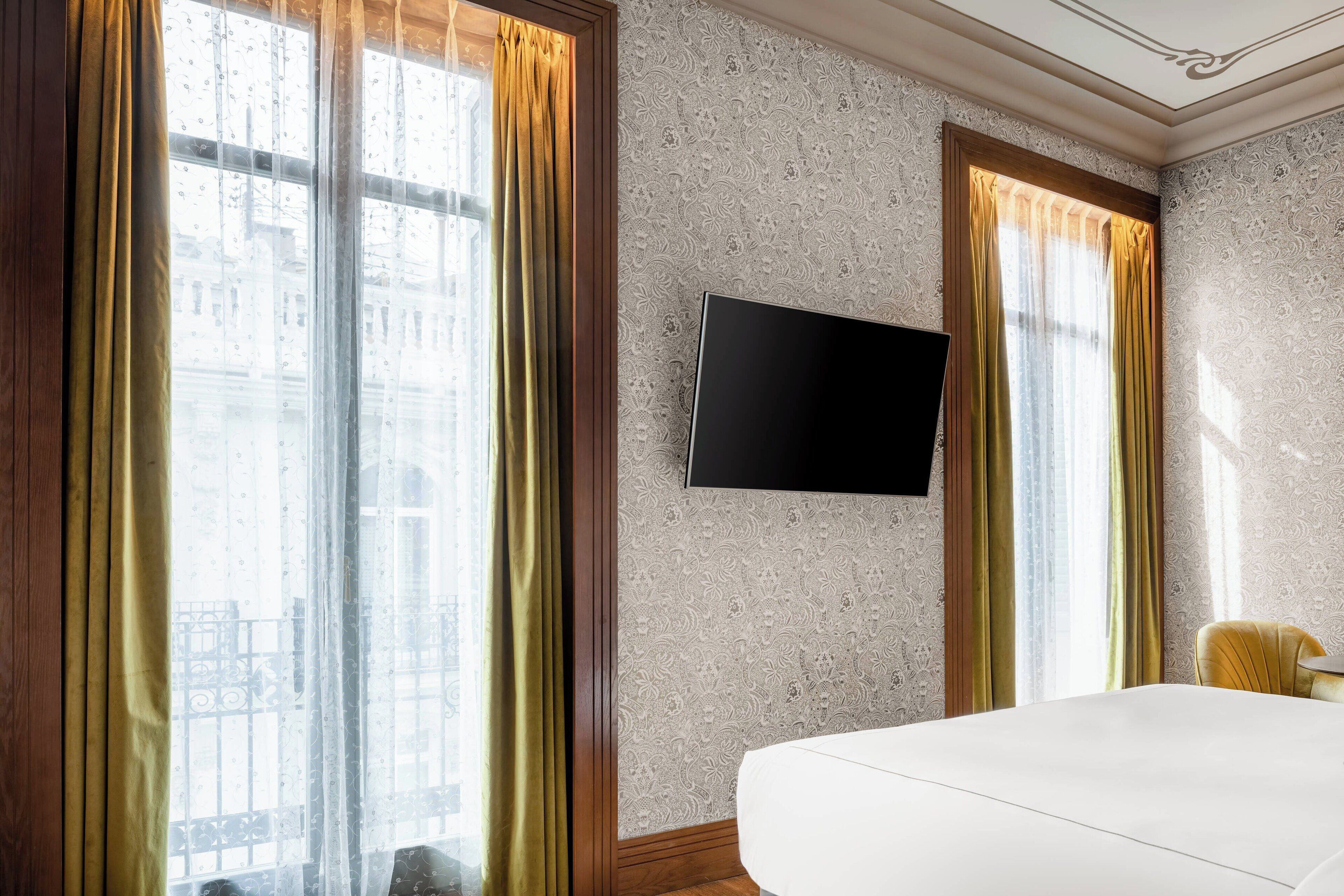 Une chambre d’hôtel dotée d’un grand lit, d’un téléviseur à écran plat fixé au mur et offrant une vue sur la ville grâce à de grandes fenêtres équipées de rideaux.