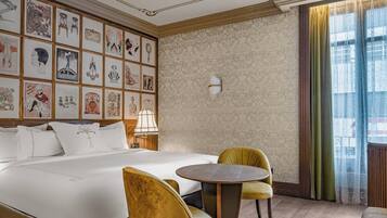 A hotel room with a large bed, a round table, and chairs, a wall with framed artwork, and a window with curtains.