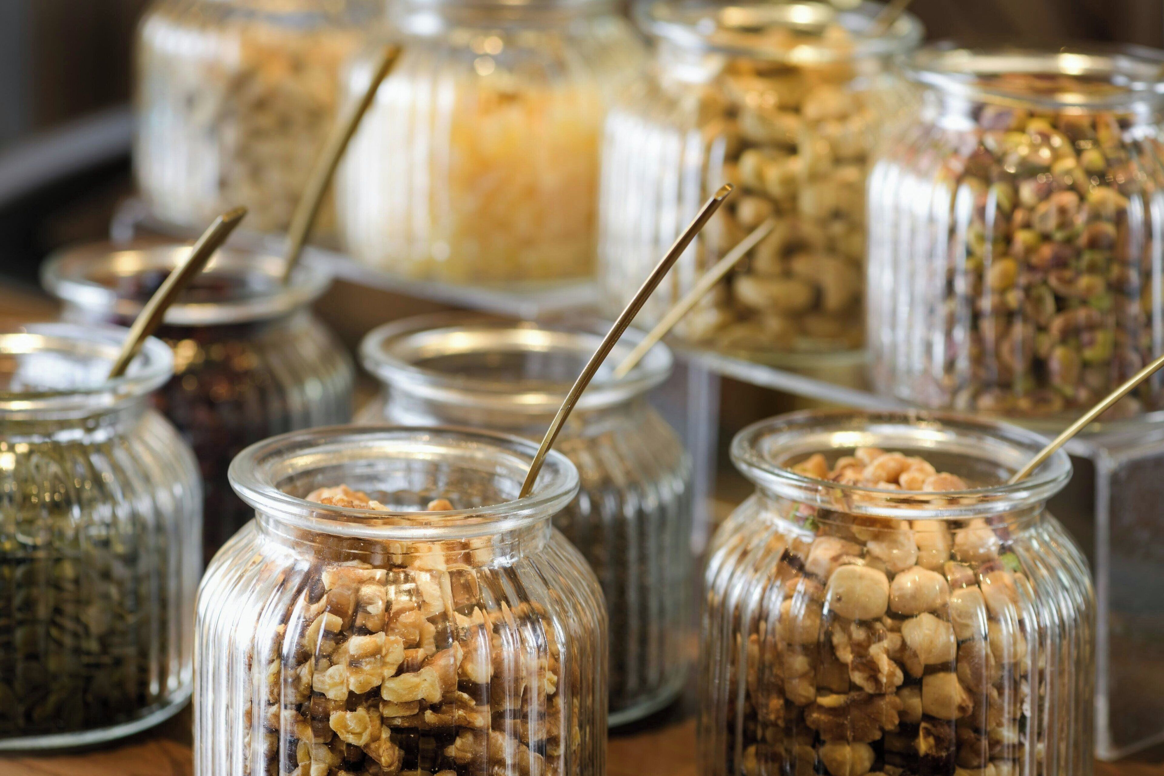 Multiple glass jars filled with various nuts and seeds, each with a metal spoon inserted.