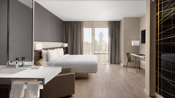 A modern hotel room with a large bed, a desk, a chair, a bathroom with a mirror and sink, and a view of the outdoors.
