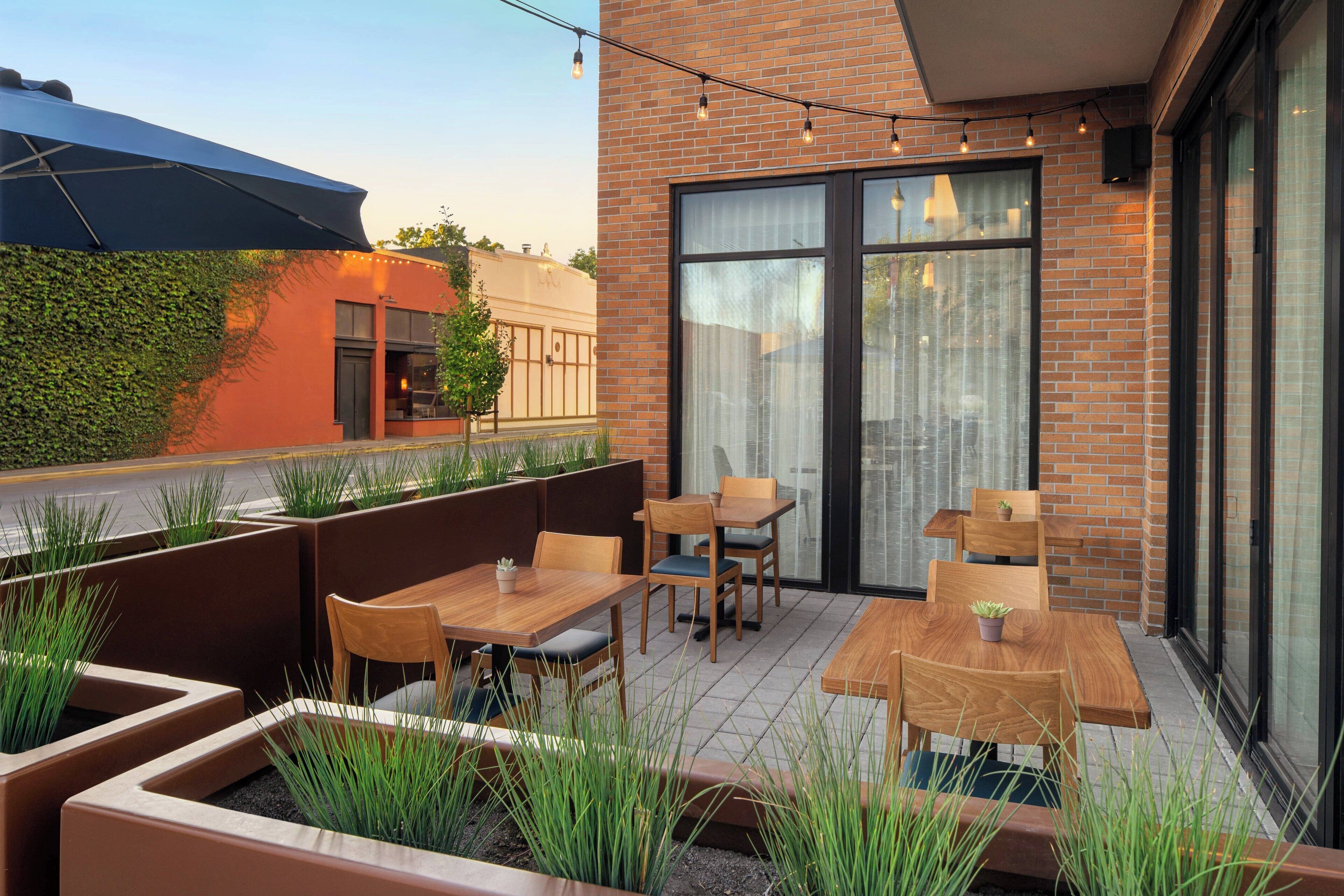 An outdoor patio with wooden tables and chairs, surrounded by planters with green plants.
