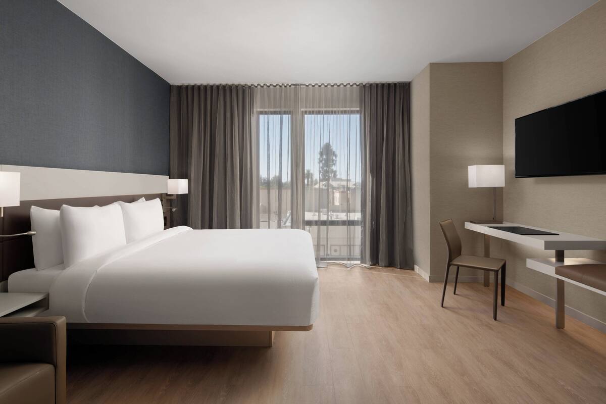 A modern hotel room with a large bed, a desk, a chair, a flat-screen TV, and a balcony with curtains.