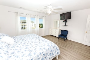 Un dormitorio con cama, cómoda, silla, ventilador de techo, televisor y ventana con cortinas.