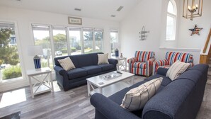 A spacious living room with a dark blue sofa, striped armchairs, and a white coffee table. Large windows offer a view of the outdoors. - Oceanside, Hot Tub, Community Amenities, Close to beach, 665, Shore Beats Work (Corolla)