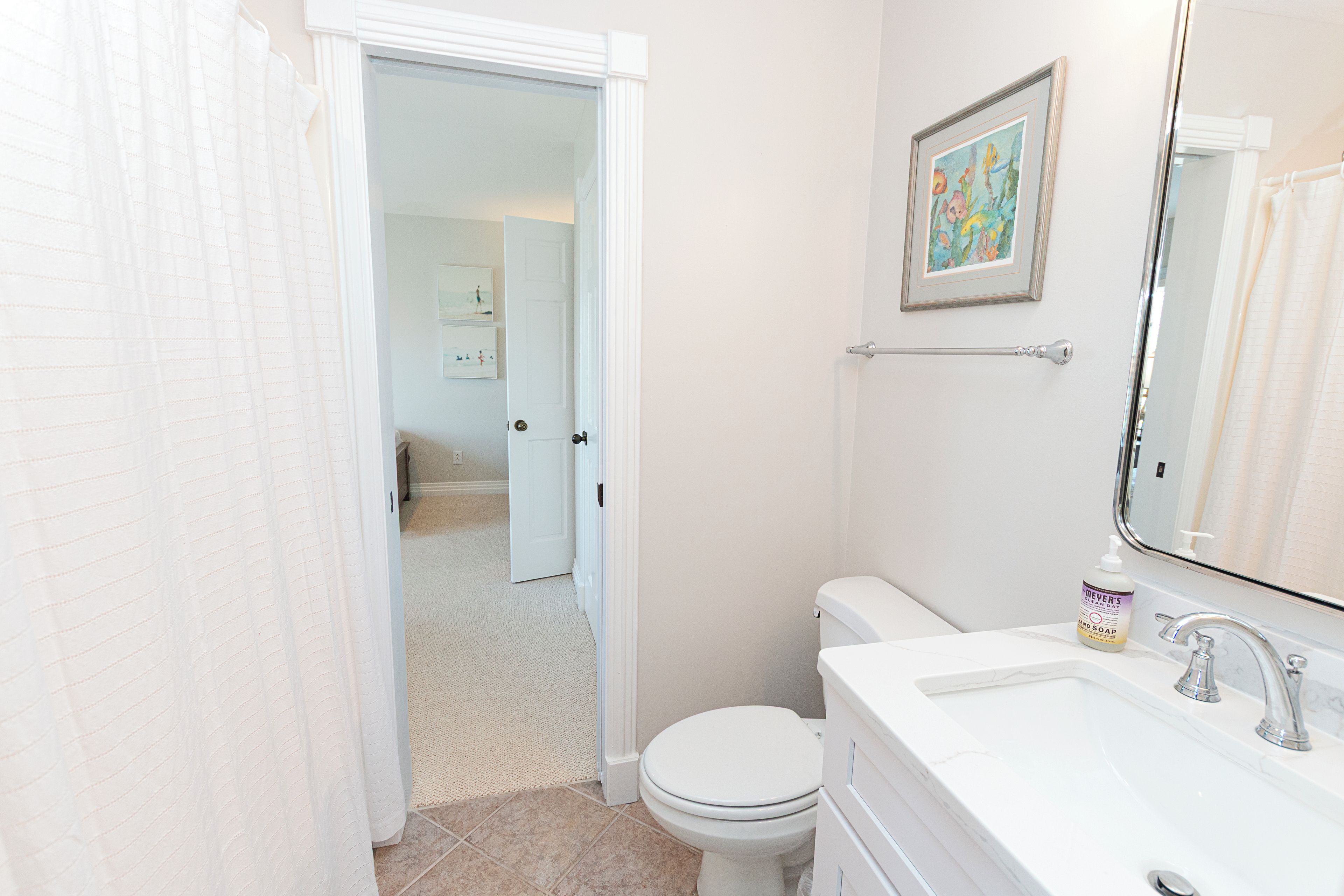 A bathroom with a toilet, sink, and a shower curtain. There is a framed picture on the wall and a bottle of hand soap on the sink.