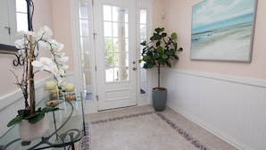Een hal met een glazen tafel, witte bloemen, een potplant en een schilderij van een strand aan de muur.