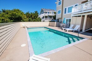 A rectangular swimming pool with a clear blue water surface, surrounded by a concrete deck. There are white lounge chairs and a small table with a blue umbrella on the right side. - Semi-Oceanfront, Private Pool, Ocean Views, Close to beach, BU76, Trading Winds (Corolla)