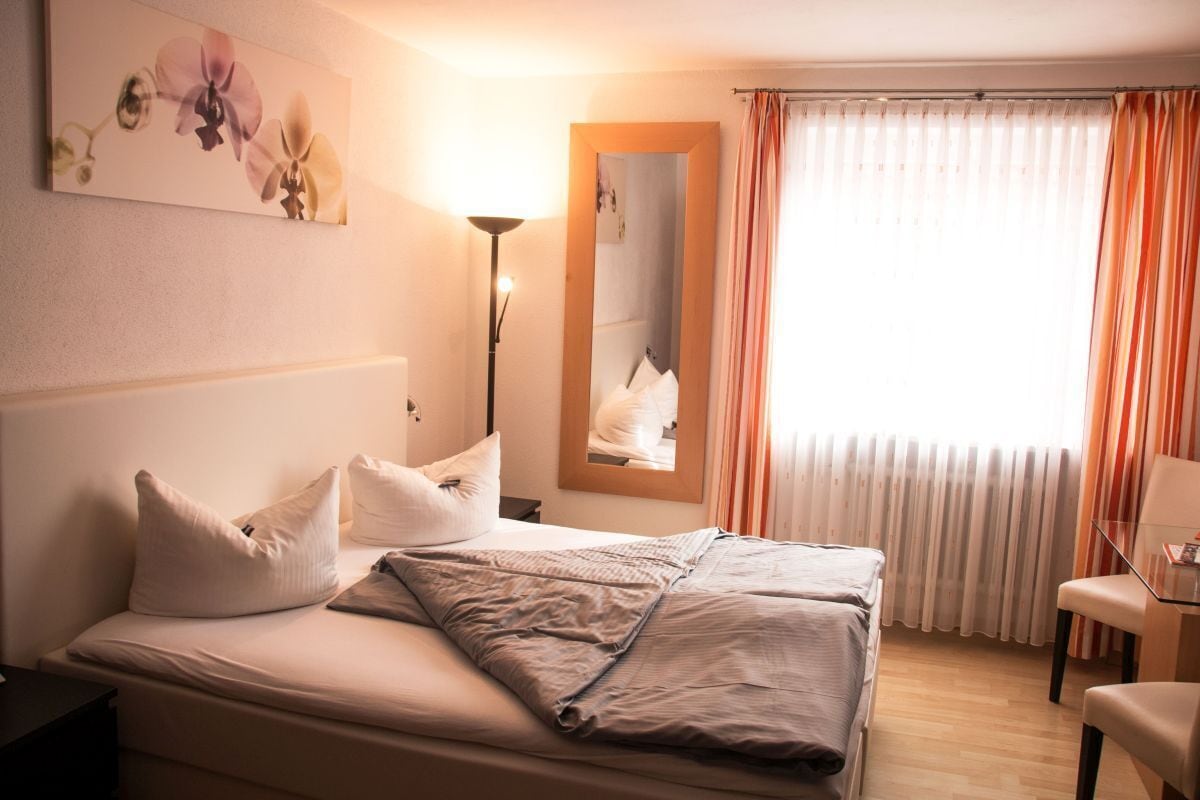 A neatly made bed with pillows, a bedside lamp, a mirror, and a window with curtains.