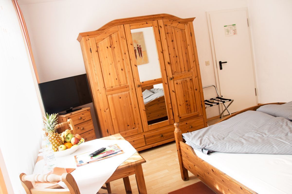 A bedroom with a wooden wardrobe, a bed with grey bedding, a wooden table with fruits and a bottle, and a TV on a stand.