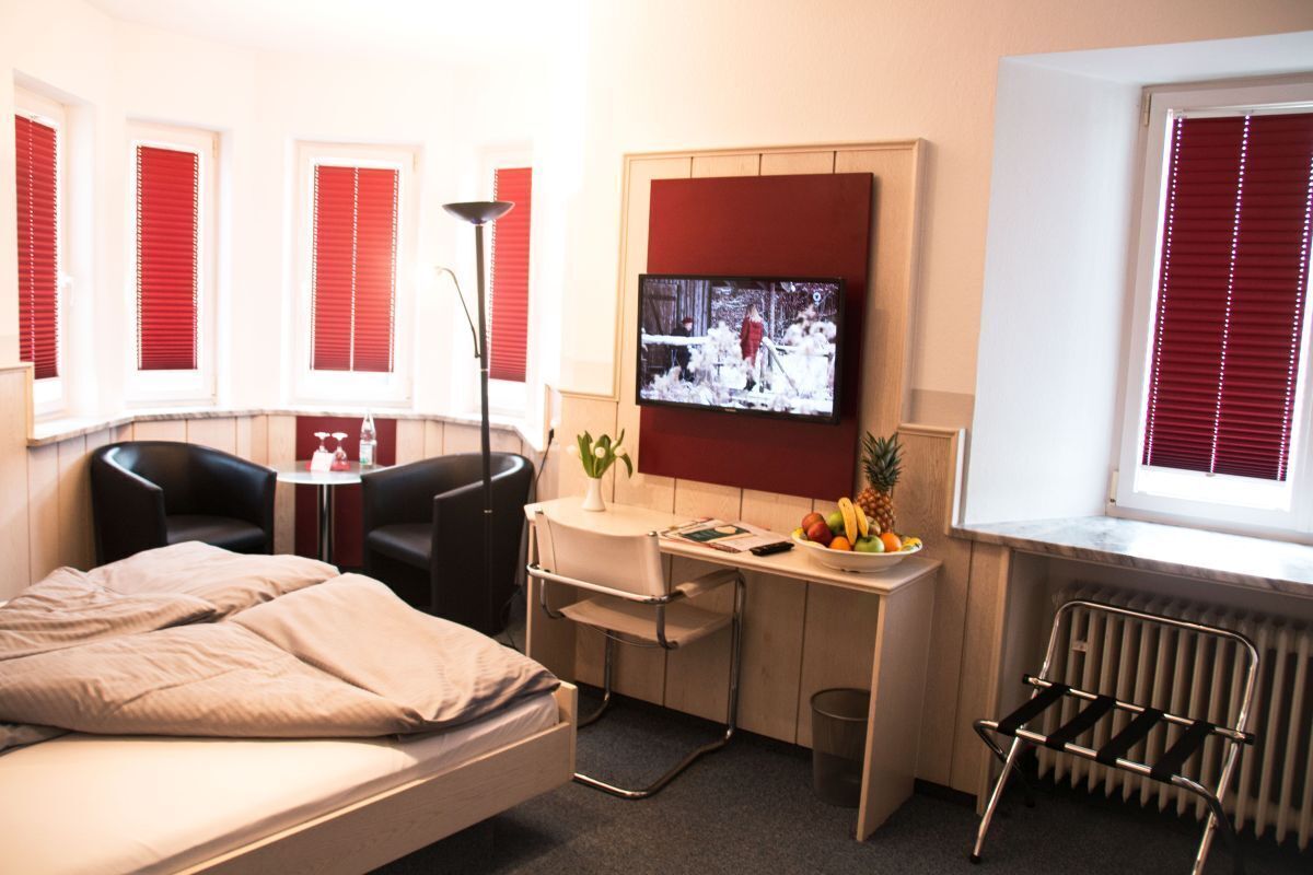 Ein kompaktes Hotelzimmer mit Bett, Schreibtisch mit Stuhl, Fernseher und Fenster mit roten Jalousien.