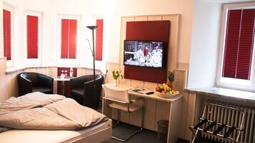 Ein kompaktes Hotelzimmer mit Bett, Schreibtisch mit Stuhl, Fernseher und Fenster mit roten Jalousien.